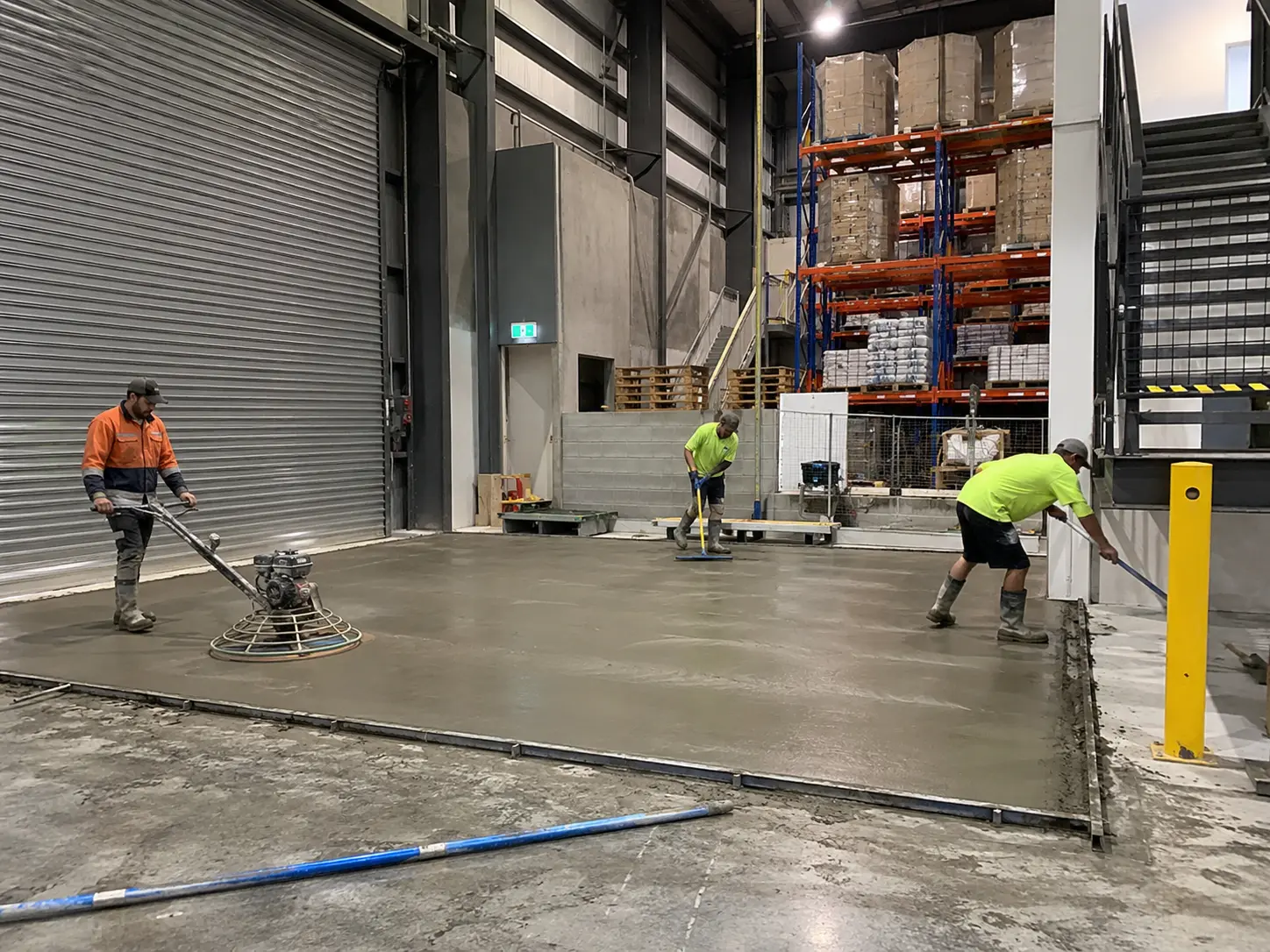 Workers power-floating a fresh concrete slab inside a working warehouse with pallet racking in the background
